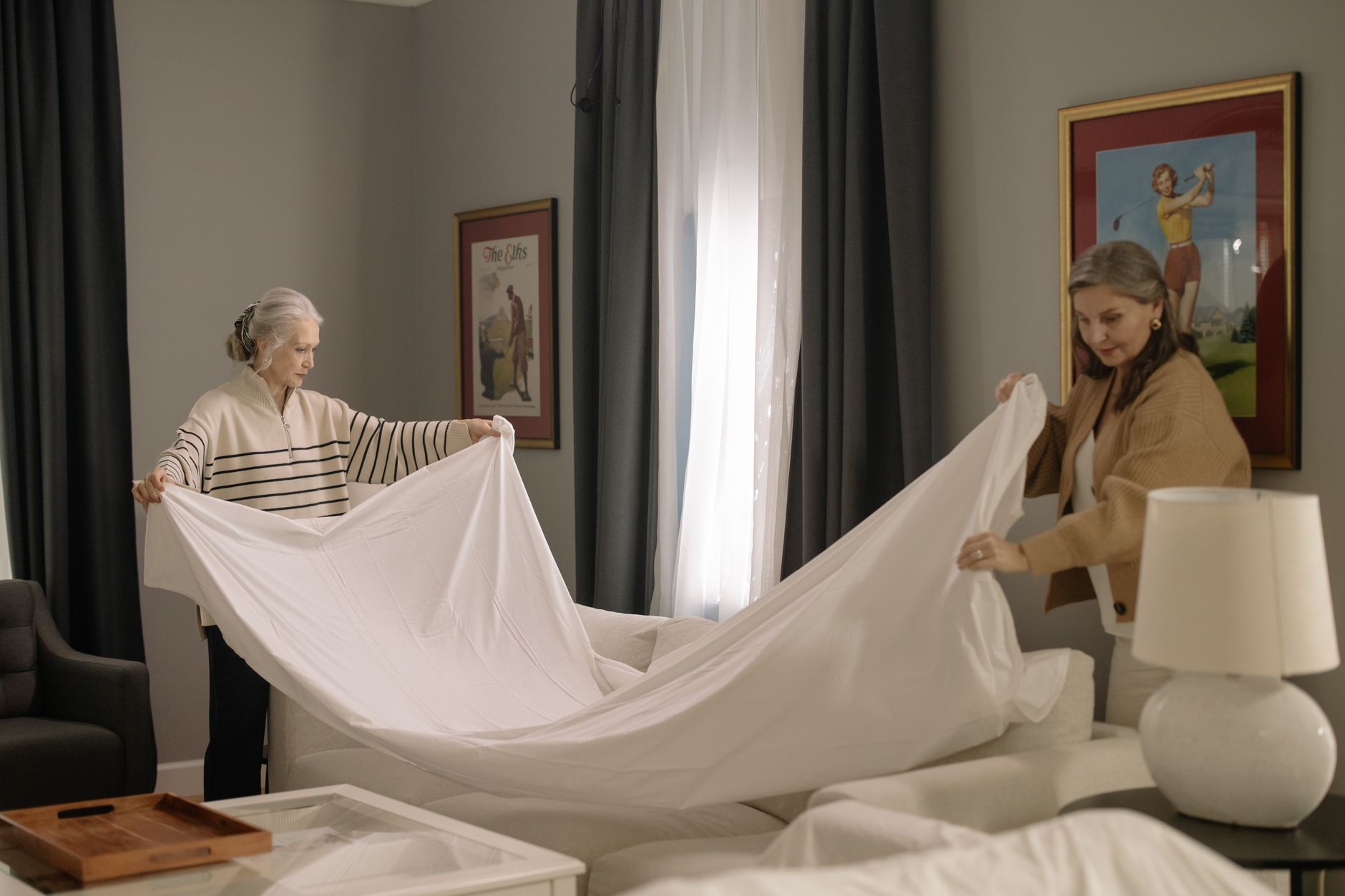 Two women spreading a white sheet over a bed