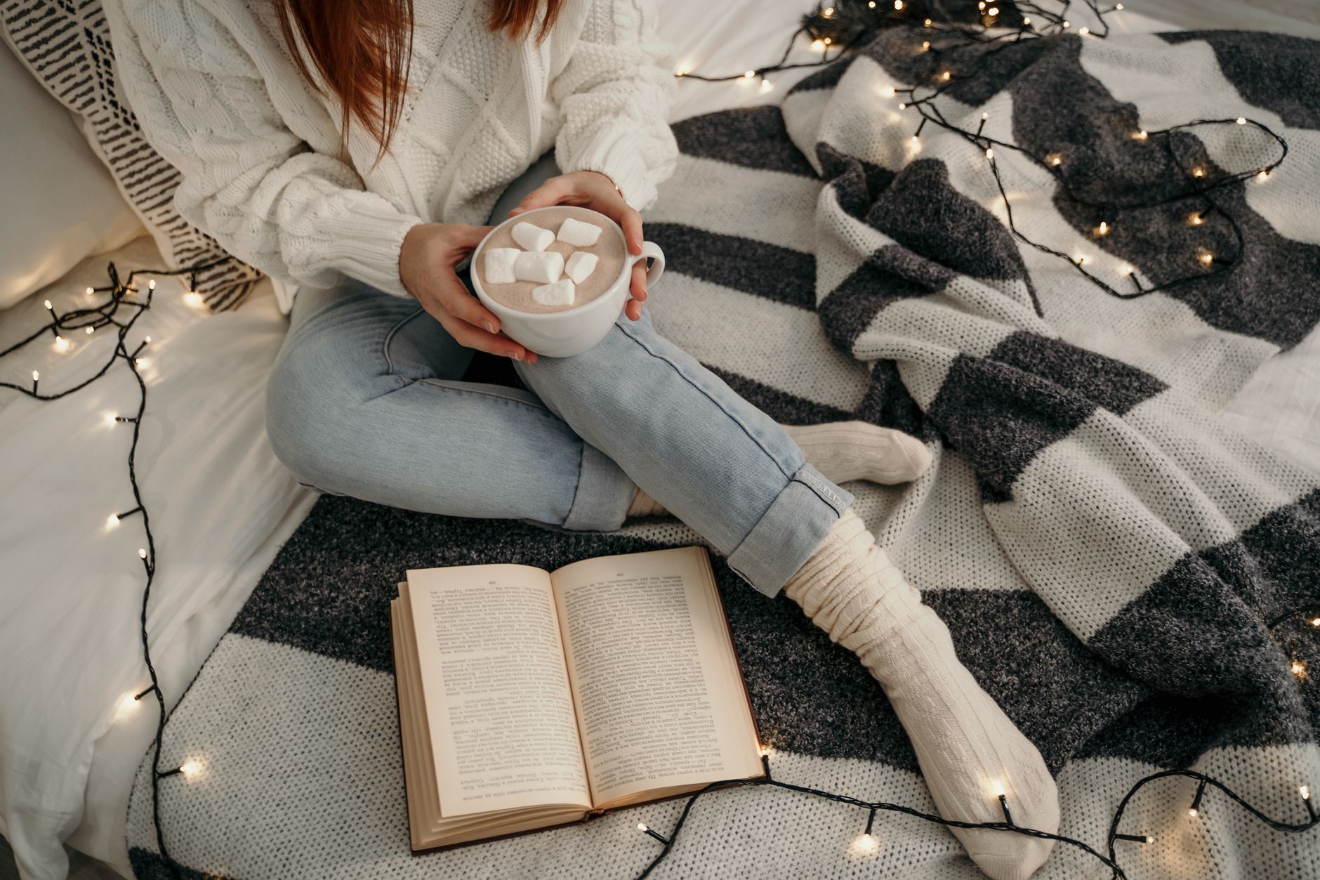 A person with cocoa and a book on a cozy blanket