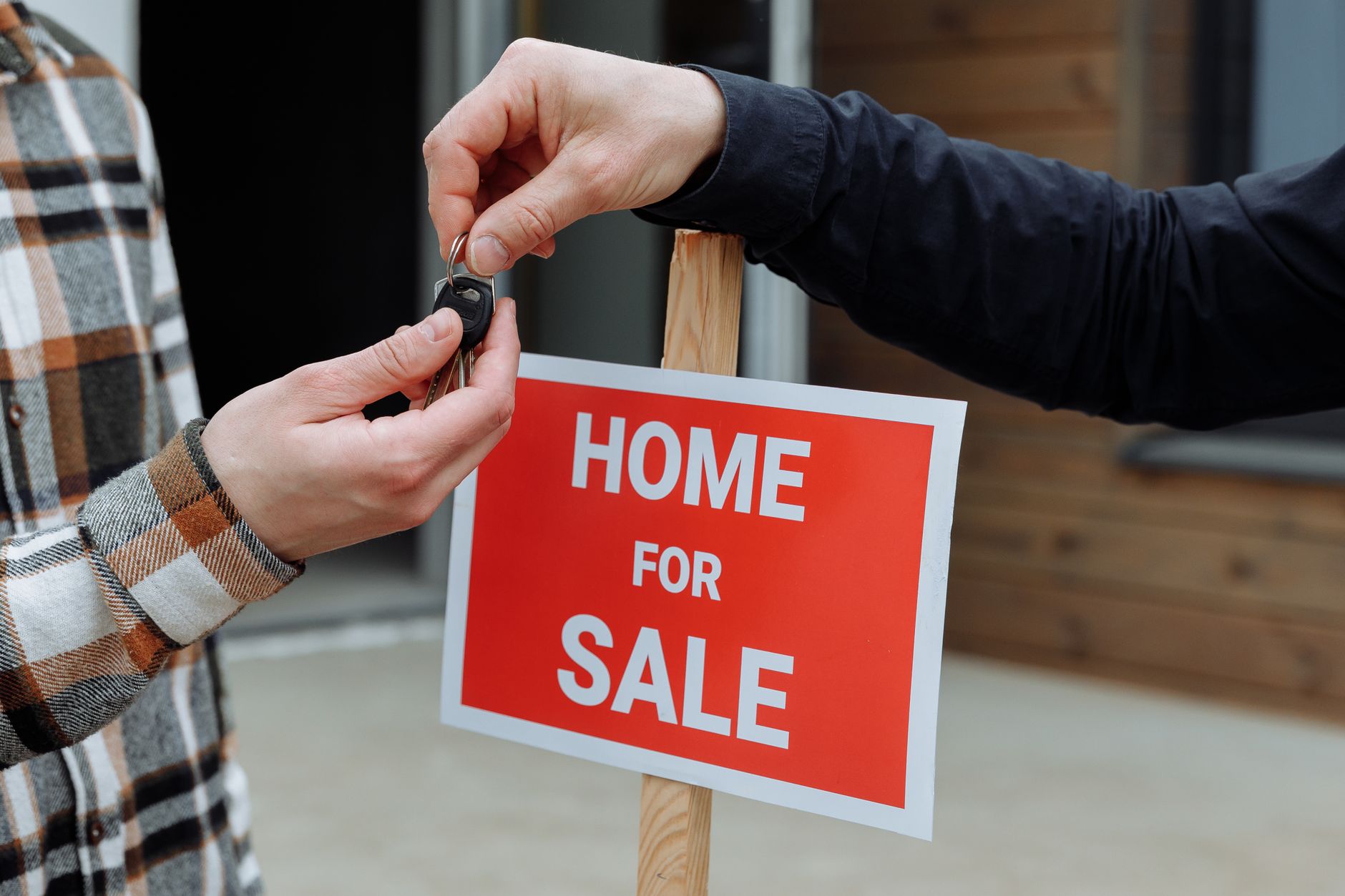 A house key being handed over in front of a for-sale sign