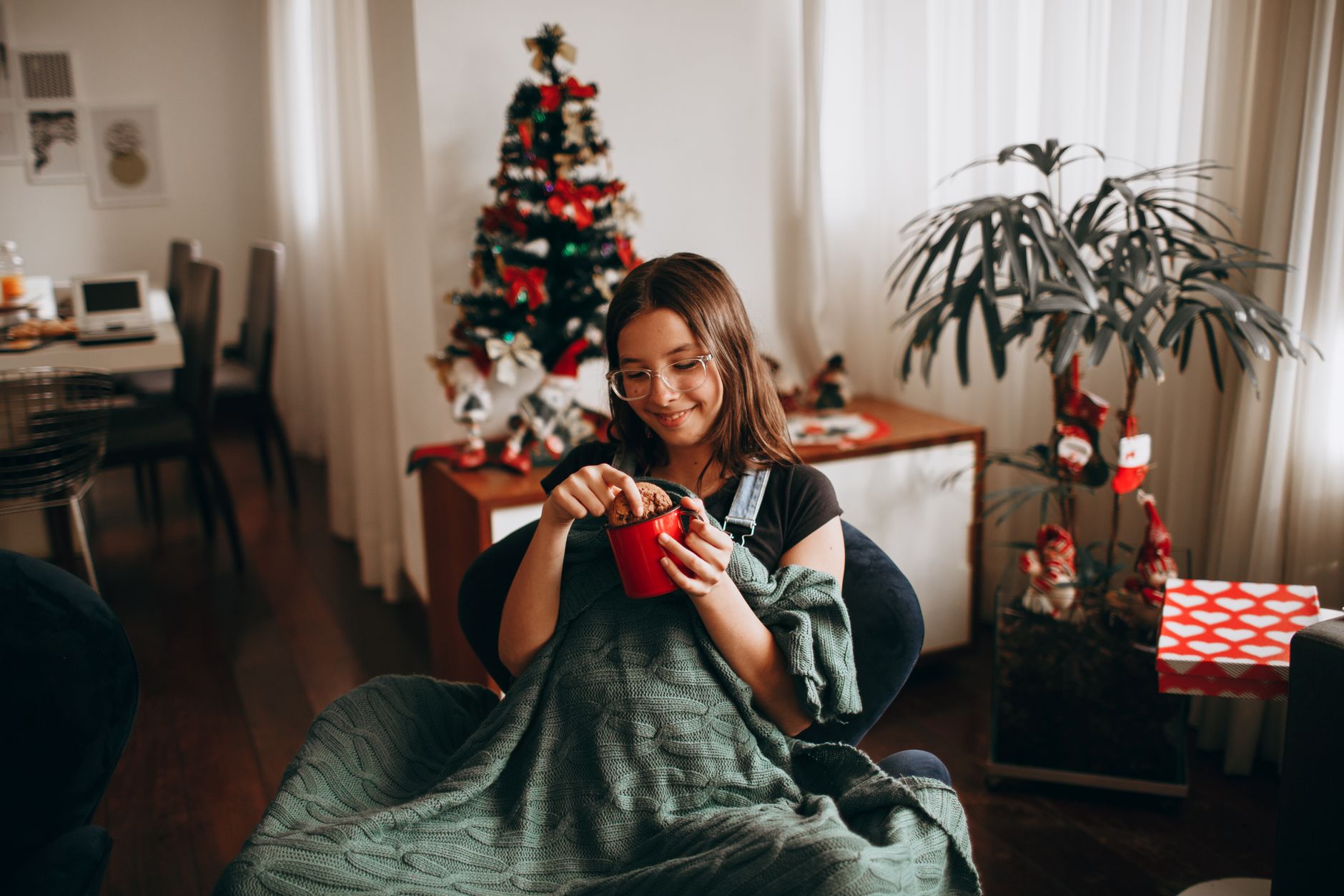 A woman with a mug under a blanket by a holiday tree